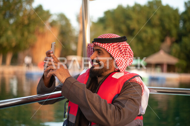 رجل عربي خليجي سعودي يرتدي سترة النجاة ، التقاط  صور تذكاريه باستخدام الهاتف المحمول ،  جولة بقارب في بحيرة مائية ، عطلة صيفية ، منتجعات ترفيهية في المملكة العربية السعودية ،  رحلات بحرية سياحية.
