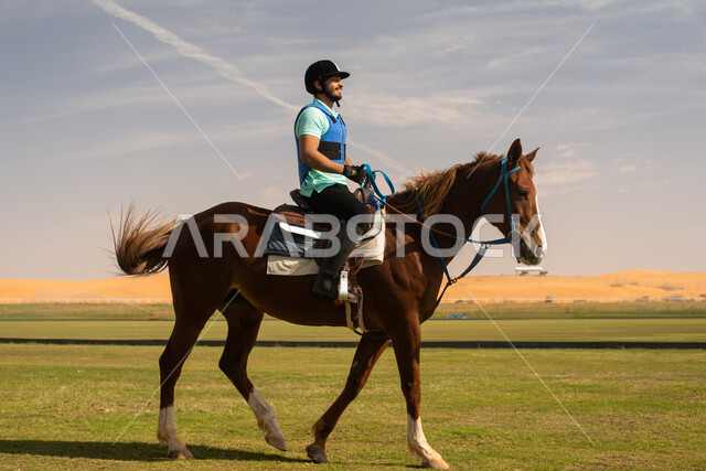 شاب عربي خليجي سعودي  يمتطي خيل عربي اصيل ويرتدي زي الفروسية, تربية الخيول في الاسطبل، هواية ركوب الخيل, حصان بني، مزرعة تدريب وترويض الخيول في السعودية ، نادي الفروسية ،انشطة ترفيهية  ممتعة .