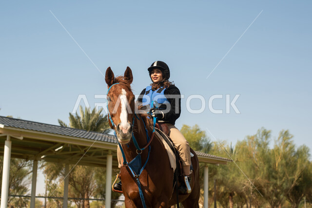 امرأة عربية  سعودية خليجية تمتطي خيل عربي أصيل وترتدي زي الفروسية, تربية الخيول في الاسطبل،  هواية ركوب الخيل, حصان بني، مزرعة تدريب وترويض الخيول في السعودية ، نادي الفروسية ،انشطة ترفيهية 