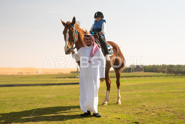 النظر الى الكاميرا ,فتى عربي خليجي سعودي مع والده يرتدي زي الفروسية ويمتطي خيل عربي أصيل, ايماءات تدل على السعادة,  حصان بني , تربية الخيول في الاسطبل, مزرعة تدريب وترويض الخيول في السعودية, نادي الفروسية, انشطة ترفيهية للاطفال .