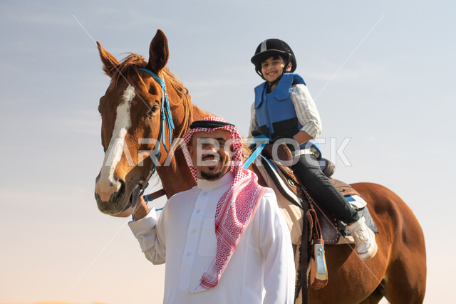صورة مقربة لفتى عربي خليجي سعودي مع والده يرتدي زي الفروسية ويمتطي خيل عربي أصيل, ايماءات تدل على السعادة,  حصان بني , تربية الخيول في الاسطبل, مزرعة تدريب وترويض الخيول في السعودية, نادي الفروسية, انشطة ترفيهية للاطفال .