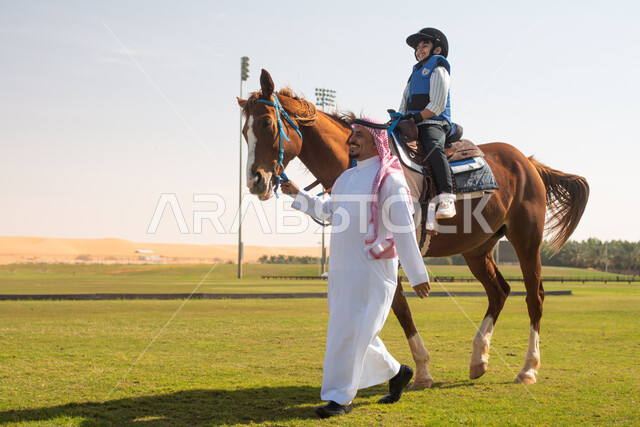 طفل عربي  سعودي خليجي مع والده يرتدي زي الفروسية ويمتطي خيل عربي أصيل, حصان بني , تربية الخيول في الاسطبل, مزرعة تدريب وترويض الخيول في السعودية, نادي الفروسية, انشطة ترفيهية للاطفال .