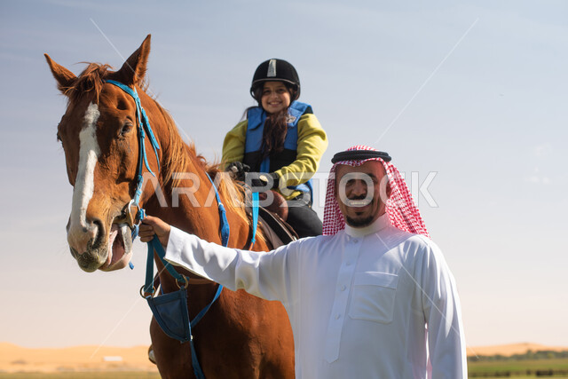صورة مقربة لفتى عربي خليجي سعودي مع والده يرتدي زي الفروسية ويمتطي خيل عربي أصيل, ايماءات تدل على السعادة,  حصان بني , تربية الخيول في الاسطبل, مزرعة تدريب وترويض الخيول في السعودية, نادي الفروسية, انشطة ترفيهية للاطفال .