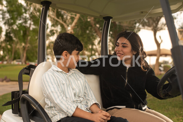 A Saudi Gulf Arab woman and her son inside an electric car specialized in transporting passengers, using a mobile phone, taking memorial photos, selfies, live broadcasting, recreational activities, golf course, golf club