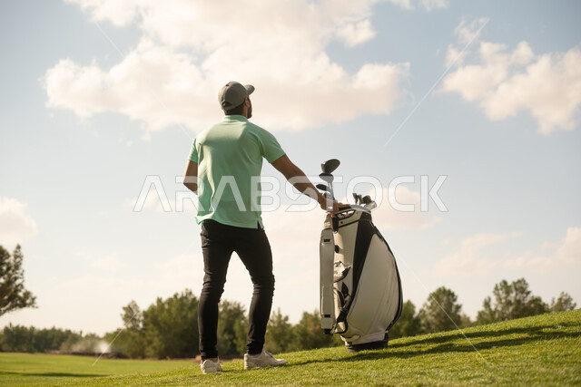 صورة من الخلف لرجل عربي خليجي سعودي يرتدي زي الكاجوال، ممارسة لعبة الغولف على مساحات واسعة خضراء، أنشطة رياضية في السعودية، رياضة لعبة الجولف، الألعاب الأولمبية الصيفية، أنشطة ترفيهية ممتعة، ملعب الجولف