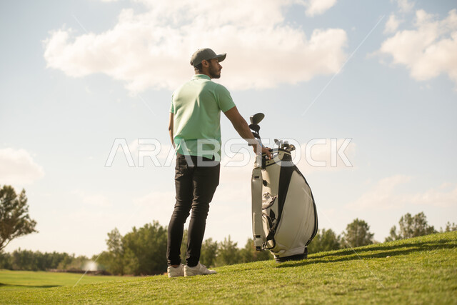 زاوية من الخلف لرجل عربي خليجي سعودي يرتدي زي الكاجوال، ممارسة لعبة الغولف على مساحات واسعة خضراء، أنشطة رياضية في السعودية، رياضة لعبة الجولف، الألعاب الأولمبية الصيفية، أنشطة ترفيهية ممتعة، ملعب الجولف