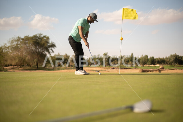 ملعب الجولف, رجل عربي خليجي سعودي يرتدي الزي الكاجوال ,ممارسة لعبة الغولف، مساحات خضراء واسعة، أنشطة رياضية في السعودية، رياضة لعبة الجولف، الألعاب الأولمبية الصيفية، أنشطة ترفيهية ممتعة، الإستمتاع باللعب