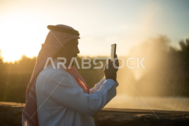 رجل عربي خليجي سعودي يقضي أوقات ممتعة في الخارج أثناء غروب الشمس ، التقاط صور تذكارية, تصوير سيلفي عبر الهاتف المحمول، منتجع ترفيهي ، الاستمتاع بالطبيعة الخضراء في الهواء الطلق، خلفية الطبيعة مع غروب الشمس .