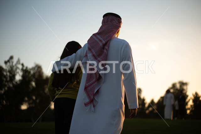 صورة من الخلف لأب عربي خليجي سعودي يقضي اوقات ممتعة مع ابنته في إحدى الحدائق, المشي والتنزه, عطلة صيفية، نزهه في الهواء الطلق، جولة في الطبيعة الخضراء، مزارع واستراحات، أنشطة ترفيهية عائلية