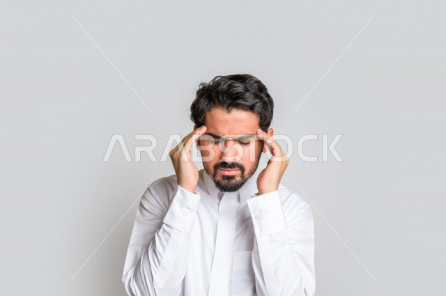 بورتريه لرجل عربي سعودي خليجي باللباس التقليدي , يقوم بإيماءات وجه ويدين تدل على الشعور بالصداع, ألم بالرأس, الإحساس بالمرض, التعب والإرهاق, الحاجه للعلاج, طلب المساعده , خلفية بيضاء .