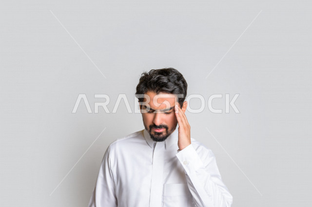 بورتريه لرجل عربي سعودي خليجي باللباس التقليدي , يقوم بإيماءات وجه ويدين تدل على الشعور بالصداع, ألم بالرأس, الإحساس بالمرض, التعب والإرهاق, الحاجه للعلاج, طلب المساعده , خلفية بيضاء .