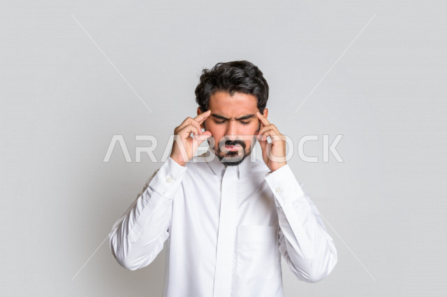 بورتريه لرجل عربي سعودي خليجي باللباس التقليدي , يقوم بإيماءات وجه ويدين تدل على الشعور بالصداع, ألم بالرأس, الإحساس بالمرض, التعب والإرهاق, الحاجه للعلاج, طلب المساعده , خلفية بيضاء .