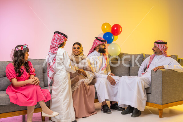 أجواء عائليه سعيده ، بورتريه لعائلة عربية خليجية سعودية تحتفل بقدوم العيد في غرفة الجلوس , تقديم العيديات للأطفال, ايماءات تدل على الفرح والسرور بالعيد السعيد, الزيارات والتجمعات الاسريه, مناسبات واعياد اسلاميه ، خلفية ملونة