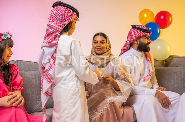 بورتريه لعائلة عربية خليجية سعودية مجتمعه في غرفة الجلوس , تقديم العيديات للأطفال, اجواء عائليه سعيده , الزيارات والتجمعات الاسريه, مناسبات واعياد اسلاميه ، خلفية ملونة