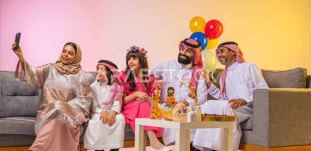 التقاط صورة سيلفي عن طريق استخدام الهاتف المحمول ، زيارات وتجمعات عائليه ، إيماءات وجه تدل على الفرح والسعادة ،بورتريه لعائلة عربية سعودية تحتفل بقدوم العيد السعيد ، الاحتفال بالعيد السعيد ، بالونات وزينة العيد ، خلفية ملونة