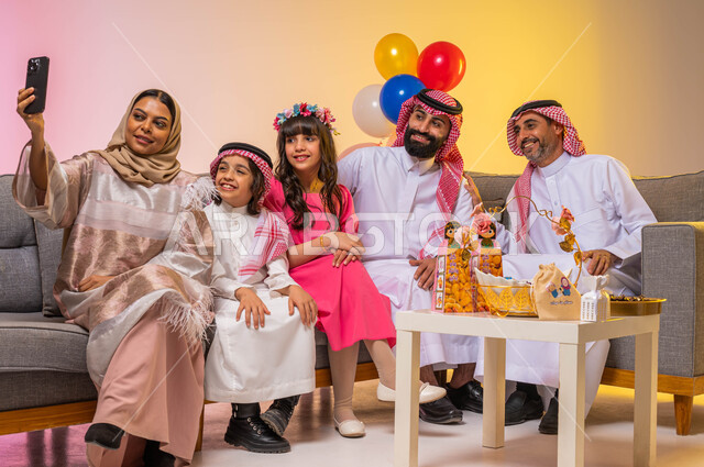 إيماءات وجه تدل على الفرح والسعادة ، زيارات وتجمعات عائليه ، بورتريه لعائلة عربية سعودية تحتفل بقدوم العيد السعيد في غرفة الجلوس ، التقاط صورة سيلفي عن طريق استخدام الهاتف المحمول ، الاحتفال بالعيد السعيد ، خلفية ملونة