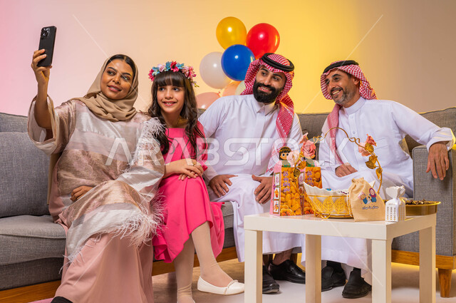 بورتريه لعائلة عربية سعودية تحتفل بقدوم العيد السعيد ، التقاط صورة سيلفي عن طريق استخدام الهاتف المحمول ، الاحتفال بالعيد السعيد ، إيماءات وجه تدل على الفرح والسعادة ، خلفية ملونة