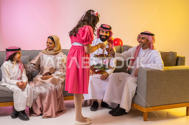 فتاة عربية خليجية سعودية تقدم حلوى العيد للضيوف ، بورتريه لعائلة عربية خليجية سعودية تحتفل بقدوم العيد في غرفة الجلوس  , مناسبات واعياد اسلاميه, زيارات وتجمعات عائليه, اكرام الضيف وحسن الاستقبال ، خلفية ملونة