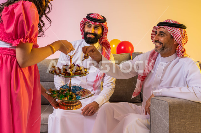بورتريه لعائلة عربية خليجية سعودية تحتفل بقدوم العيد في غرفة الجلوس ، فتاة عربية خليجية سعودية  تقدم حلوى العيد للضيوف , مناسبات واعياد اسلاميه, زيارات وتجمعات عائليه, اكرام الضيف وحسن الاستقبال ، خلفية ملونة