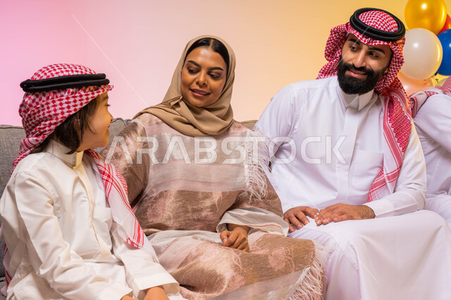 بورتريه لعائلة عربية خليجية سعودية  تحتفل بقدوم العيد في غرفة الجلوس  ، إيماءات وجه تدل على الفرح والسعادة ، الاحتفال بقدوم العيد ،أجواء العيد في السعودية ، خلفية ملونة