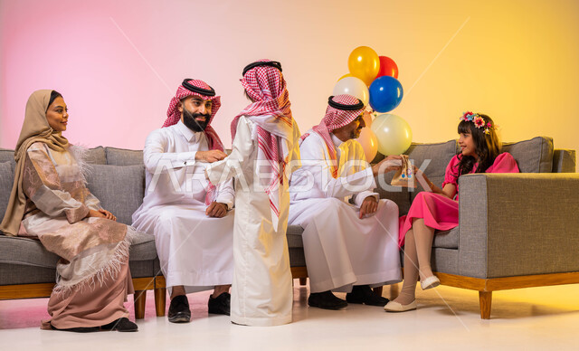 إيماءات وجه تدل على الفرح والسعادة ، بورتريه لعائلة عربية خليجية سعودية تحتفل بقدوم العيد في غرفة الجلوس  ،  تقديم الحلوى و قرقيعان العيد ،أجواء العيد في السعودية ،  خلفية ملونة