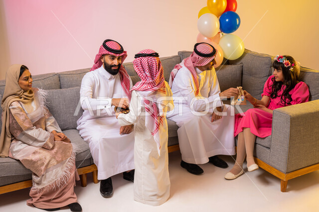 التهنئة بقدوم العيد ، تقديم الحلوى و قرقيعان العيد ، ارتداء اللباس السعودي التقليدي ، أجواء العيد في المملكة العربية السعودية ، بورتريه لعائلة عربية خليجية سعودية تحتفل بالعيد في غرفة المعيشة ، خلفية ملونة