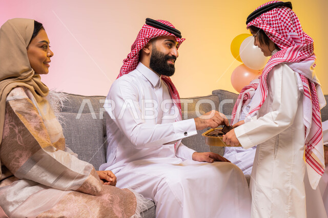 تقديم الحلوى وضيافة العيد ، أجواء العيد في المملكة العربية السعودية ، بورتريه لعائلة عربية خليجية سعودية تحتفل بالعيد في غرفة الجلوس ، ارتداء اللباس السعودي التقليدي ، التهنئة بقدوم العيد ، خلفية ملونة