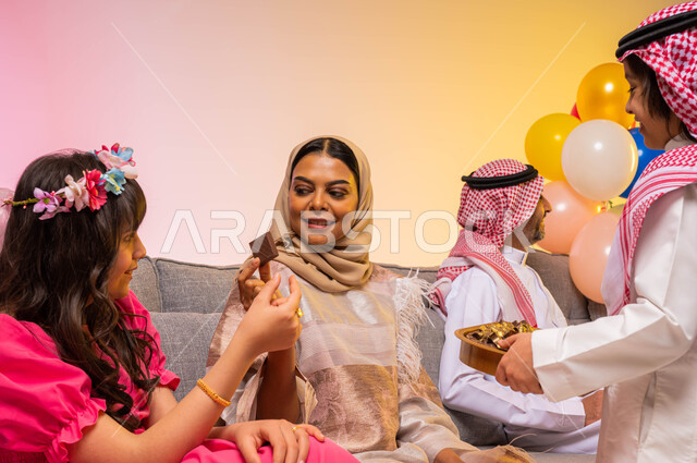 بورتريه لعائلة عربية خليجية سعودية تحتفل بالعيد في غرفة المعيشة ، ارتداء اللباس السعودي التقليدي ، تبادل أطراف الحديث ، تقديم حلوى وكعك العيد ، أجواء العيد في السعودية ، خلفية ملونة