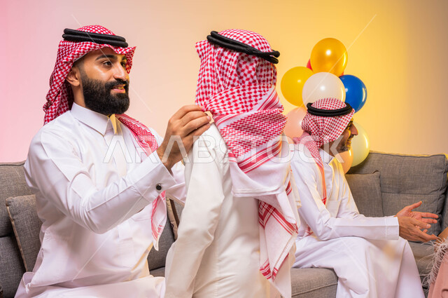 بورتريه لرجل عربي سعودي خليجي يرتدي الزي السعودي التقليدي يقوم بمساعدة ابنه على ارتداء ملابس العيد, التهيؤ والتحضير للعيد السعيد, أجواء عائليه سعيده, أعياد ومناسبات اسلاميه ، خلفية ملونة