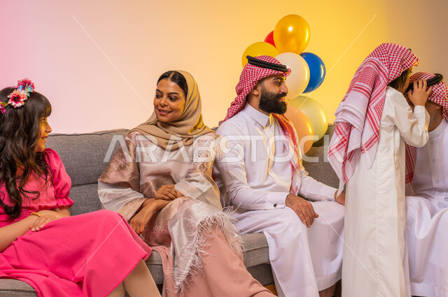 بورتريه لعائلة عربية خليجية سعودية تحتفل بقدوم العيد السعيد في غرفة المعيشة ، الزيارات العائلية والتجمعات الأسرية ، ولد عربي سعودي خليجي يقوم بتقبيل رأس جده, التهنئه بالعيد السعيد, ايماءات تدل على الفرح والسرور ، خلفية ملونة