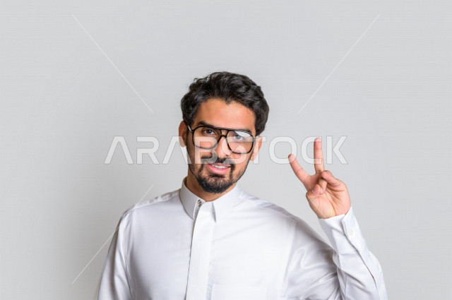 بورتريه لرجل عربي سعودي خليجي يرتدي الثوب السعودي, ايماءات وجه ويدين تدل على الفوز والإنتصار, تحقيق الاهداف, إنجاز المهام , علامة النصر والقوه , الشعور بالسعاده والرضى , خلفية بيضاء .