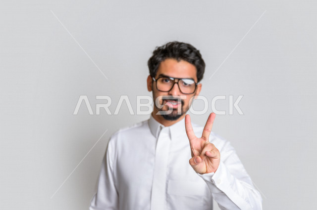 بورتريه لرجل عربي سعودي خليجي يرتدي الثوب السعودي, ايماءات وجه ويدين تدل على الفوز والإنتصار, تحقيق الاهداف, إنجاز المهام , علامة النصر والقوه , الشعور بالسعاده والرضى , خلفية بيضاء .