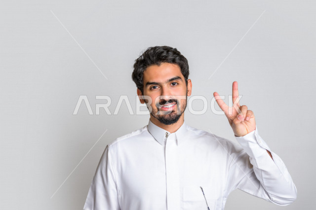 بورتريه لرجل عربي سعودي خليجي يرتدي الثوب السعودي, ايماءات وجه ويدين تدل على الفوز والإنتصار, تحقيق الاهداف, إنجاز المهام , علامة النصر والقوه , الشعور بالسعاده والرضى , خلفية بيضاء .