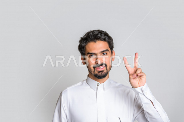 بورتريه لرجل عربي سعودي خليجي يرتدي الثوب السعودي, ايماءات وجه ويدين تدل على الفوز والإنتصار, تحقيق الاهداف, إنجاز المهام , علامة النصر والقوه , الشعور بالسعاده والرضى , خلفية بيضاء .
