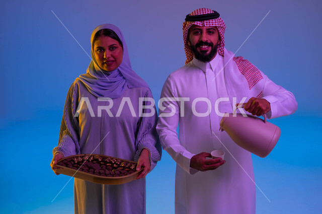 زوجان عربيان خليجيان سعوديان يحملان ضيافة العيد، رجل يصب القهوة في الفنجان، امرأة تحمل طبق حلوى العيد، اجواء العائلة السعودية في العيد، بهجة العيد، عادات العيد في المجتمع السعودي، العيد بين الأهل، الاحتفال بالعيد السعيد، اعياد ومناسبات