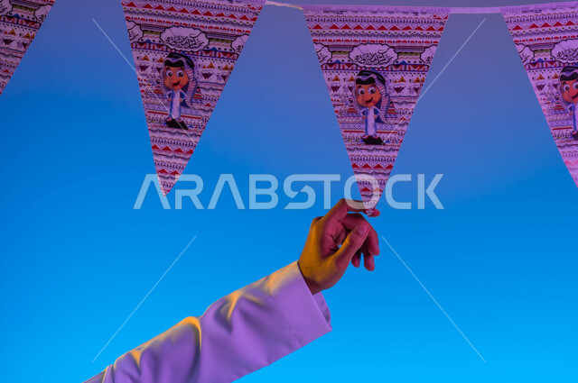 صورة مقربة ليد رجل عربي خليجي سعودي يمسك زينة العيد، بهجه وفرحة العيد، استقبال العيد، الاحتفال بالعيد السعيد، أعياد ومناسبات، خلفية ملونة 