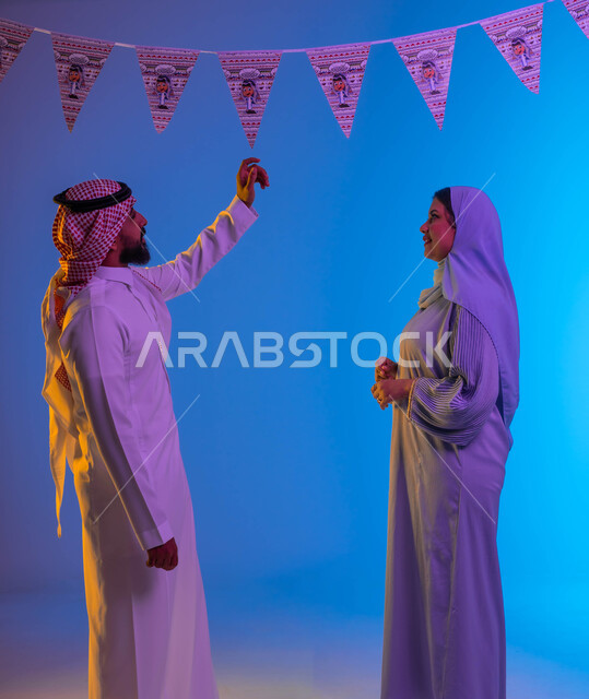رجل يمسك زينة العيد، اجواء العائلة السعودية في العيد، زوجان عربيان خليجيان سعوديان، بهجه وفرحة العيد، استقبال العيد، العيد بين الأهل، تجمع العائلة، الاحتفال بالعيد السعيد، أعياد ومناسبات، خلفية ملونة 