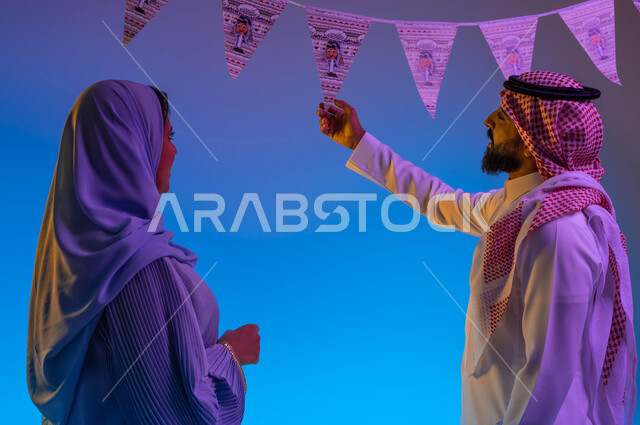 زوجان عربيان خليجيان سعوديان، اجواء العائلة السعودية في العيد، رجل يمسك زينة العيد، بهجه وفرحة العيد، استقبال العيد، العيد بين الأهل، تجمع العائلة، الاحتفال بالعيد السعيد، أعياد ومناسبات، خلفية ملونة 