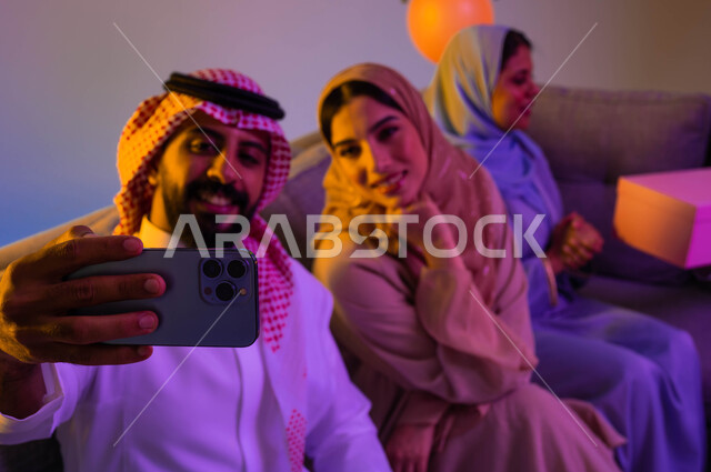 رجل وامرأتان عربيان خليجيان سعوديان يلتقطون صور السيلفي التذكارية، استخدام الهاتف المحمول، بث مباشر، اجواء العائلة السعودية في العيد، العيد بين الاهل، بهجة وفرحة العيد، اللمة العائلية، العيد في المجتمع السعودي، زيارات العيد، الاحتفال بالعيد السعيد