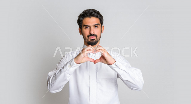 بورتريه لرجل عربي سعودي خليجي يرتدي الثوب السعودي, إشارات يدين على شكل قلب حب , إيماءات وجه ويدين تدل على الشعور بالحب, الوقوع في الغرام, السعاده والطمأنينه, المحبه والهيام, عيد الحب , خلفية بيضاء