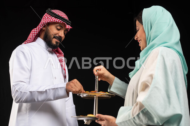 بورتريه تقديم الضيافة وحلوى العيد ، زوجان عربيان خليجيان سعوديان ، إيماءات وجه تدل على الفرح والسعادة  ، الاحتفال بقدوم العيد ،أجواء العيد في السعودية ، تقديم حلوى العيد ، خلفية سوداء
