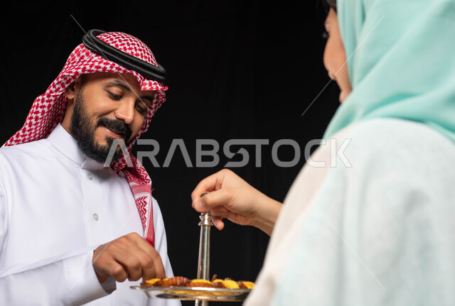تقديم الضيافة وحلوى العيد ، زوجان عربيان خليجيان سعوديان ، إيماءات وجه تدل على الفرح والسعادة  ، الاحتفال بقدوم العيد ،أجواء العيد في السعودية ، تقديم حلوى العيد ، خلفية سوداء