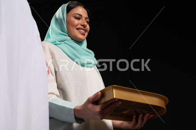 بورتريه مقرب لشابة عربيه سعوديه خليجيه تقوم بتقديم حلوى العيد للضيوف, مناسبات واعياد اسلاميه, زيارات وتجمعات عائليه, اكرام الضيف وحسن الاستقبال, خلفيه سوداء
