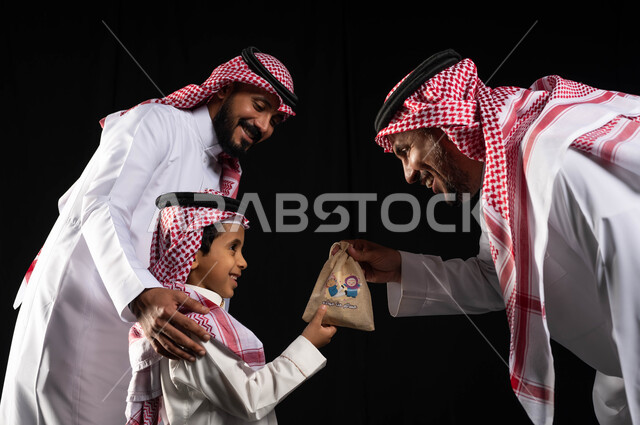 إيماءات وجه تدل على الفرح والسعادة ، تقديم العيدية للأطفال ، بورتريه لعائلة عربية خليجية سعودية ترتدي ملابس الزي التقليدي السعودي ، الاحتفال بقدوم العيد ،أجواء العيد في السعودية ، زيارات العيد ، خلفية سوداء