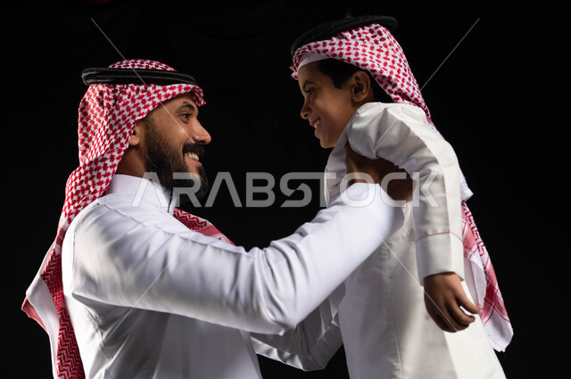 بورتريه لأب عربي سعودي خليجي يرتدي الزي السعودي التقليدي يقوم برفع ابنه للاعلى, قضاء أوقات ممتعه مع الأبناء, ايماءات تدل على الحب والاهتمام بالاولاد, ارتداء الزي السعودي التقليدي في الأعياد والمناسبات, أجواء عائليه سعيده, عيد مبارك, خلفيه سوداء