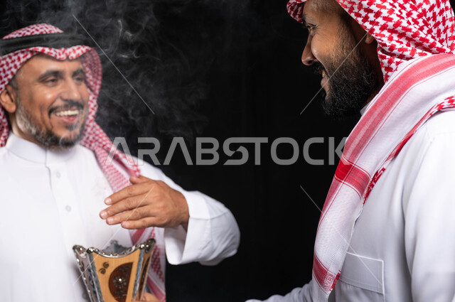 التطيب والتعطر ، استخدام خشب العود ، زيارات العيد ، حامل البخور( المبخرة ) ، بورتريه لرجلان خليجيان سعوديان مبتسمان داخل المنزل يحتفلان بالعيد ، أجواء العيد في السعودية ، خلفية سوداء