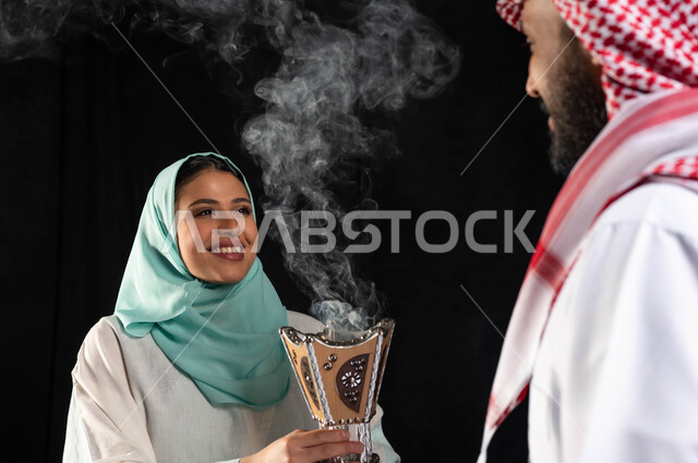 رجل عربي خليجي سعودي يرتدي الزي السعودي ، بورتريه لأمرأة عربية سعودية خليجية محجبة ترتدي العباءه الخليجية ، امرأة تحمل حامل البخور( المبخرة ) ، استخدام خشب العود في المناسبات الاجتماعية والدينية ، خلفية سوداء