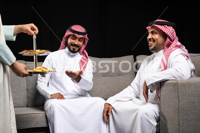 تقديم الضيافة وحلوى العيد لشابان عربيان خليجيان سعوديان يرتديان الثوب السعودي التقليدي ، الفرح بقدوم العيد ، التجمعات الأسرية في العيد ، الاحتفال بالأعياد الإسلامية ، خلفية سوداء