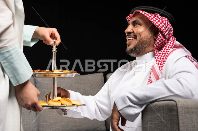 بورتريه مقرب لرجل عربي سعودي خليجي يرتدي الزي السعودي التقليدي , تقديم حلوى وكعك العيد , حسن استقبال واكرام الضيوف, ايماءات تدل على الفرح والسعاده, الزيارات و التجمعات الاسريه, أعياد ومناسبات, التهنئه بقدوم العيد, خلفيه سوداء