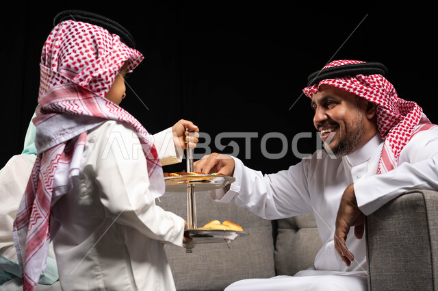 Portrait of a Saudi Arabian Gulf man wearing traditional Saudi dress, offering sweets and Eid cakes, welcoming and honoring guests, gestures indicating joy and happiness, family visits and gatherings, holidays and occasions, congratulations on the advent 